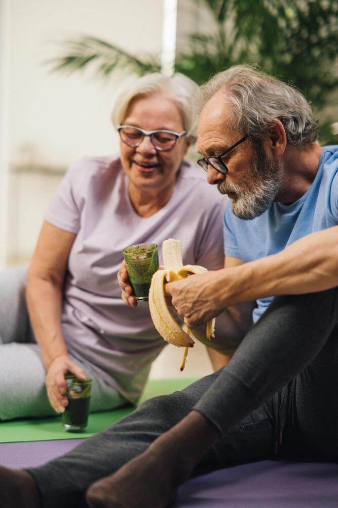 An older couple eating a banana while sitting on a yoga mat.